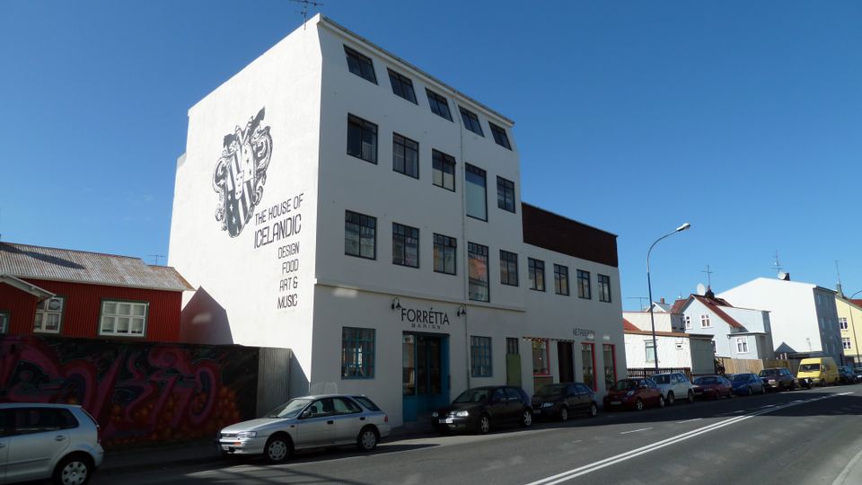 A white multi-story building with text painted on the facade sits next to a street lined with parked cars.