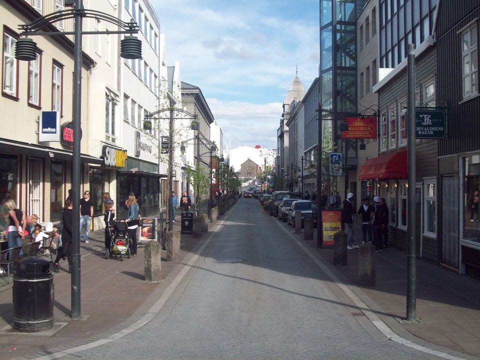 Pedestrians walk along a paved city street with shops and storefronts on either side.