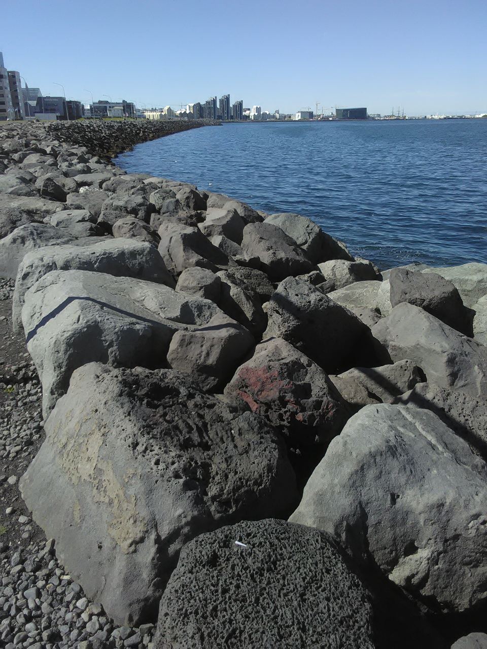 A rocky breakwater along the water with the distant Reykjavik skyline under a clear blue sky.