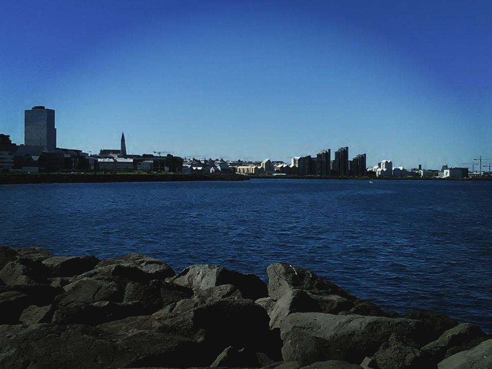 View of the Reykjavik city skyline across blue water from a rocky shore.