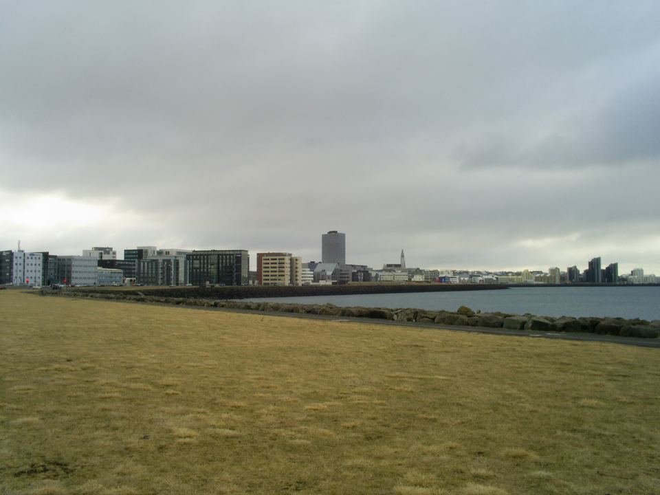 Wide view of the Reykjavik coastal skyline beyond a grassy field under cloudy skies.