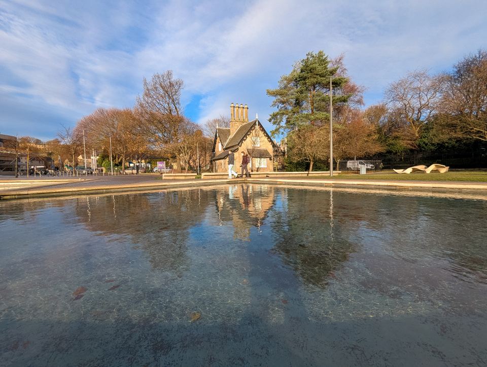 A wide reflection pool with a stone cottage building and bare trees in a UK park.