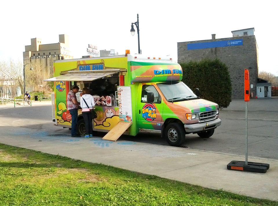 A colorful food truck parked on a sunny paved lot with patrons ordering at the window.