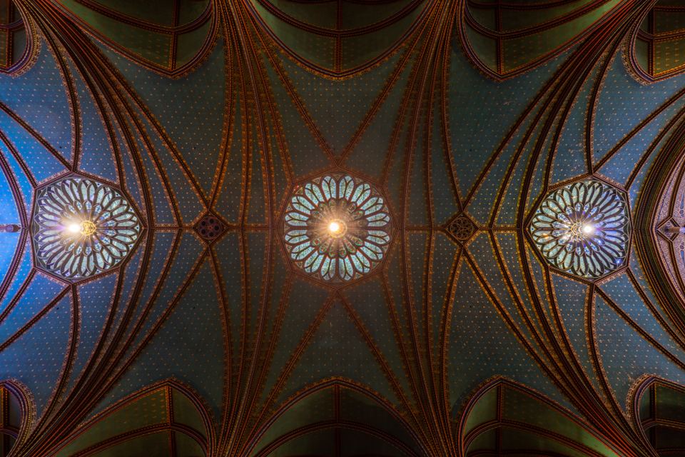 Direct upward view of blue vaulted church ceiling adorned with golden stars and stained glass