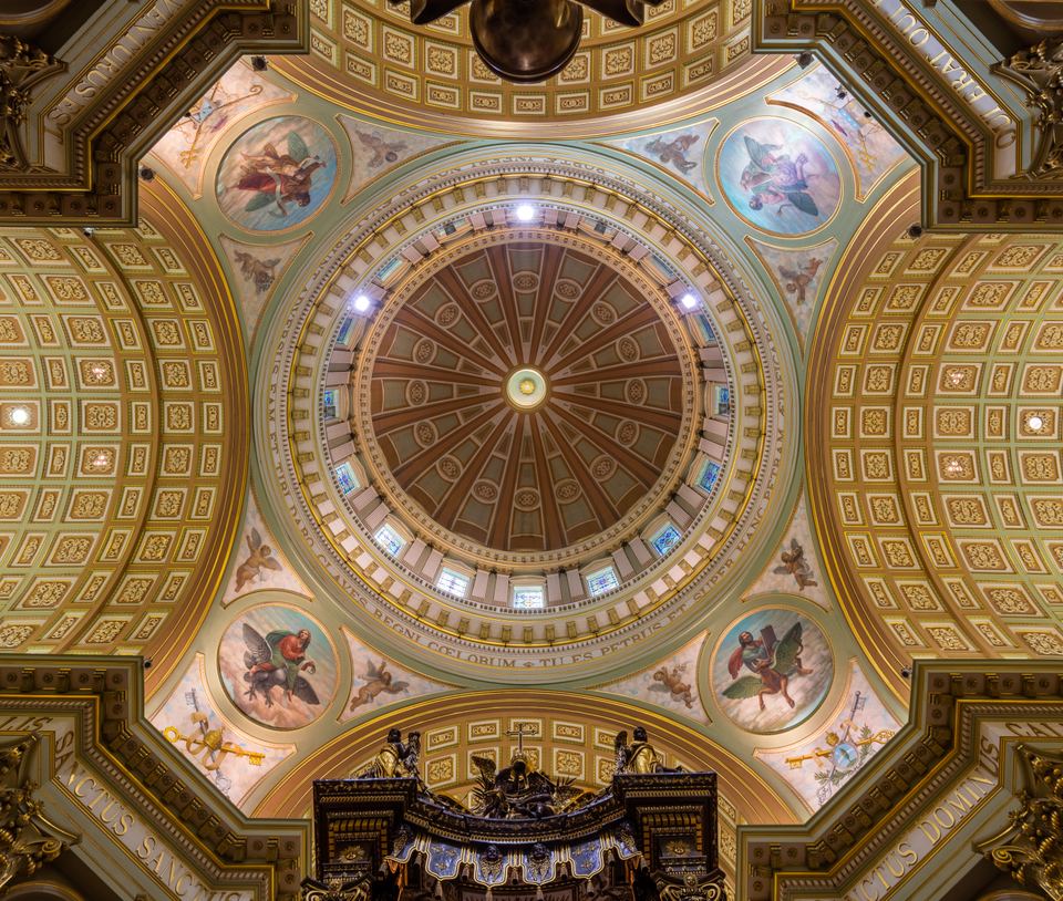 Directly upward view of a circular decorated cathedral dome with gilded details and paintings.