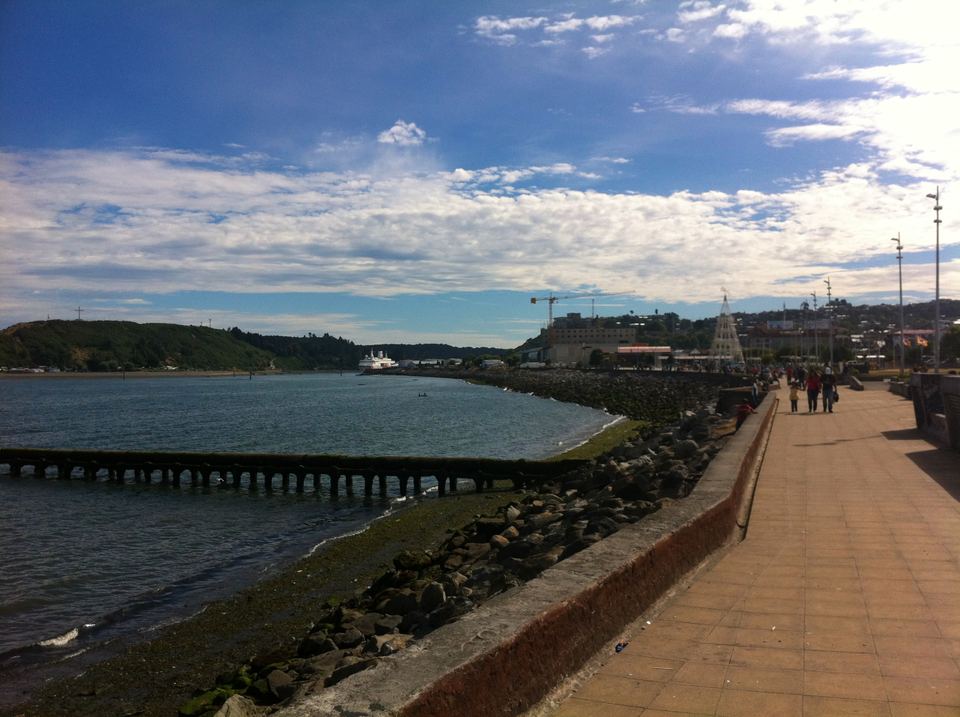 A wide paved waterfront path curving along a rocky shoreline under a blue sky