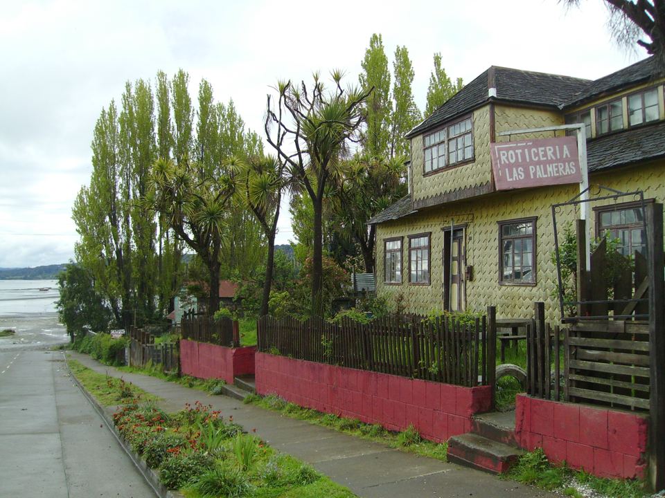 A shingled wooden house beside a street with trees and water in the background.