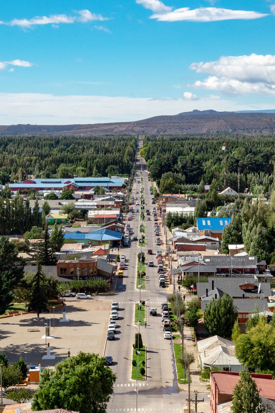 Aerial view of a long straight road running through a rural town with green trees and hills in the background.
