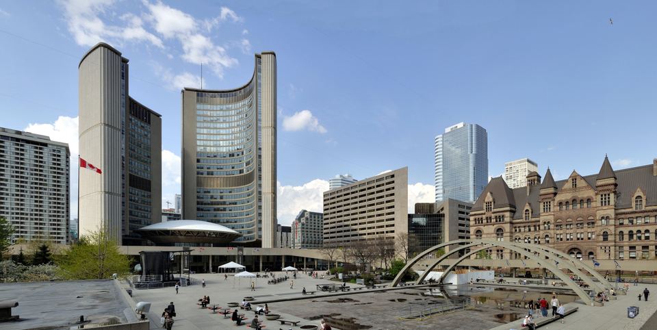 Wide view of a large public plaza with modern curved towers and a historic stone building.
