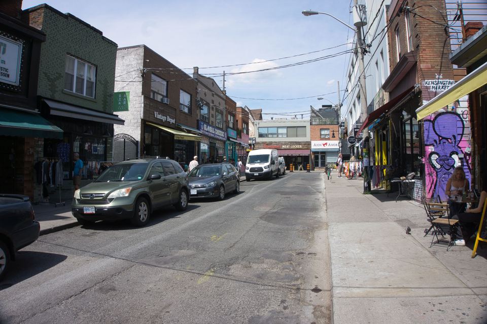 A sunny street scene in Kensington Market with boutique shops, parked cars, and outdoor seating.