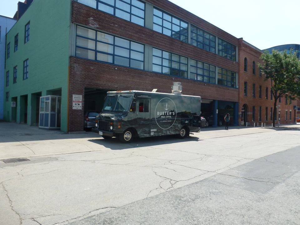 A dark food truck parked on a paved street beside a brick building under bright sunlight.
