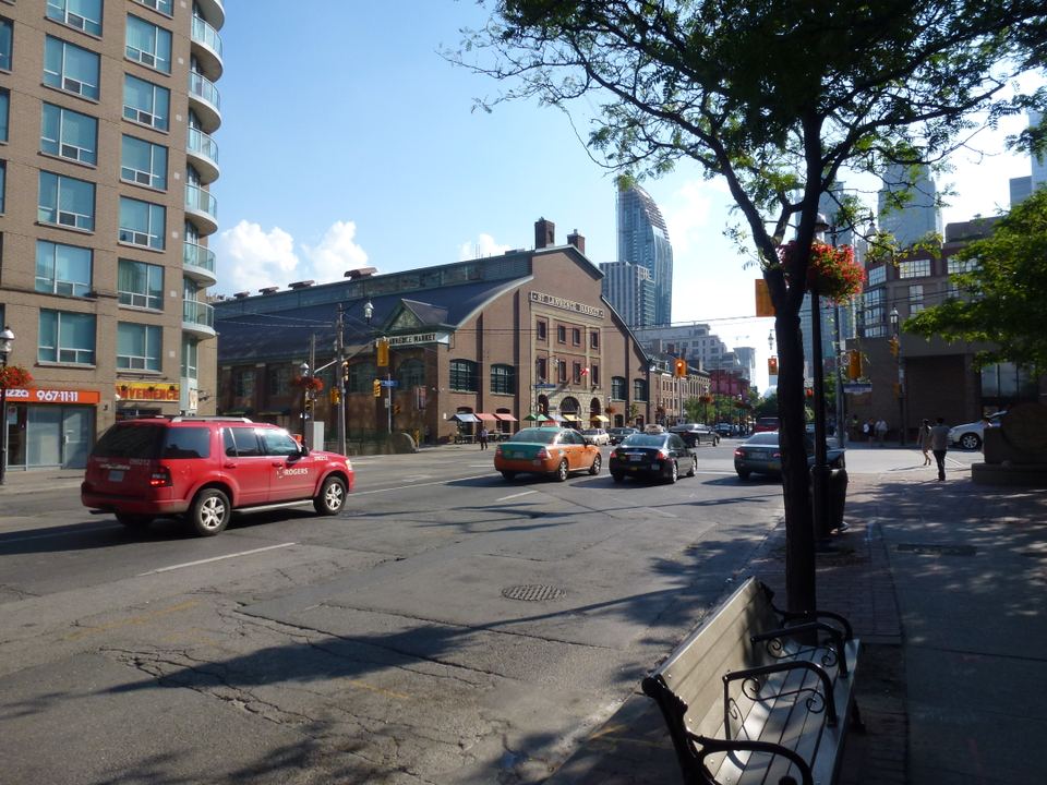 A city street scene featuring cars driving past the brick St. Lawrence Market building under a clear blue sky.