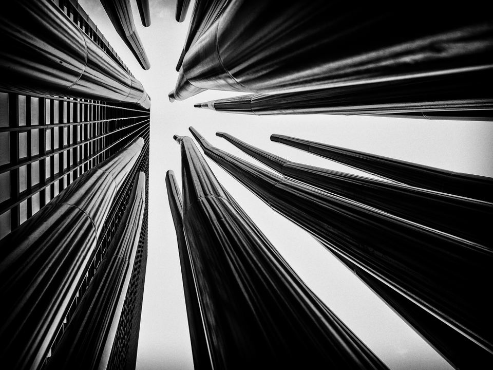 A black and white low-angle shot looking directly up at tall metallic architectural tubes against high-rise buildings.