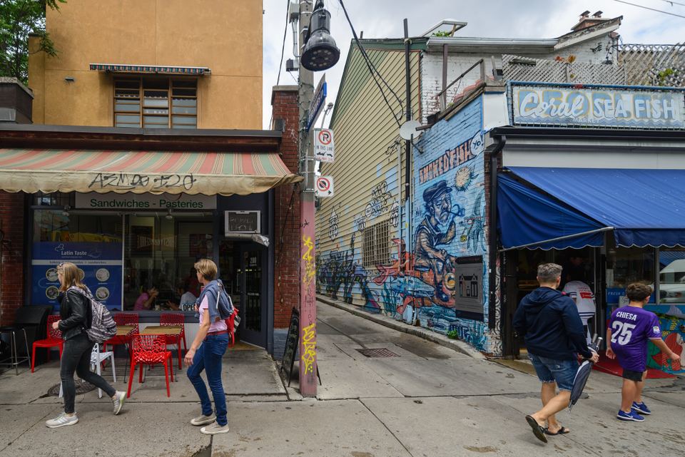 Pedestrians walking along a street lined with small shops and street art murals.