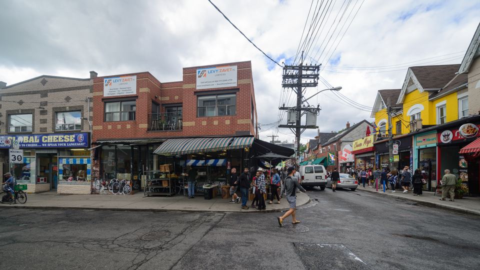 People walking on a Toronto street past brick market shops under a cloudy sky