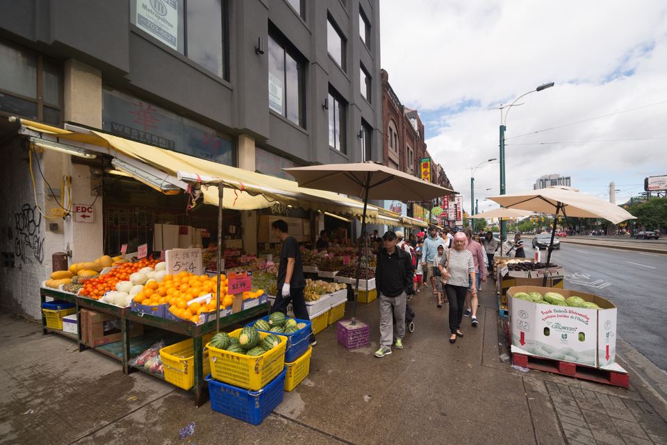 Shoppers walking past crates of vibrant oranges and watermelons on a sidewalk market display