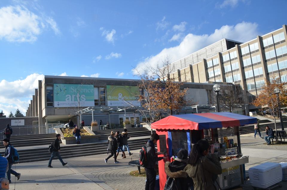 A red food cart parked in front of a modern building with students walking by.