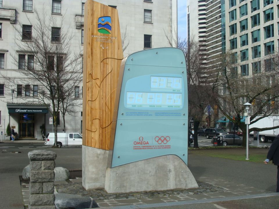 A large wooden and glass Olympic countdown clock standing on a downtown city street.