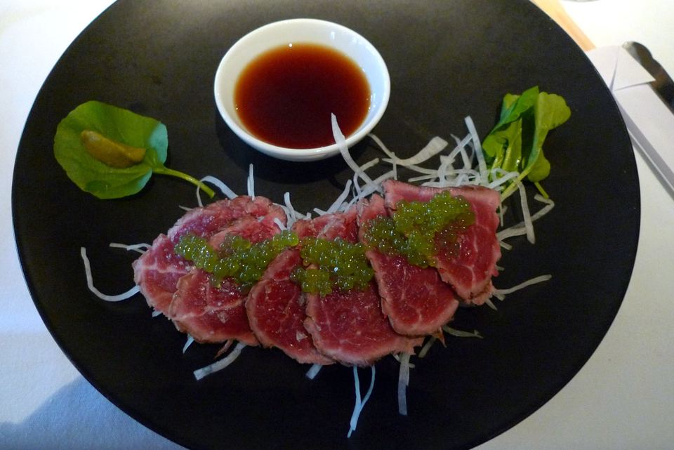 A black plate featuring arranged beef tataki slices, green garnishes, and a small bowl of dipping sauce.
