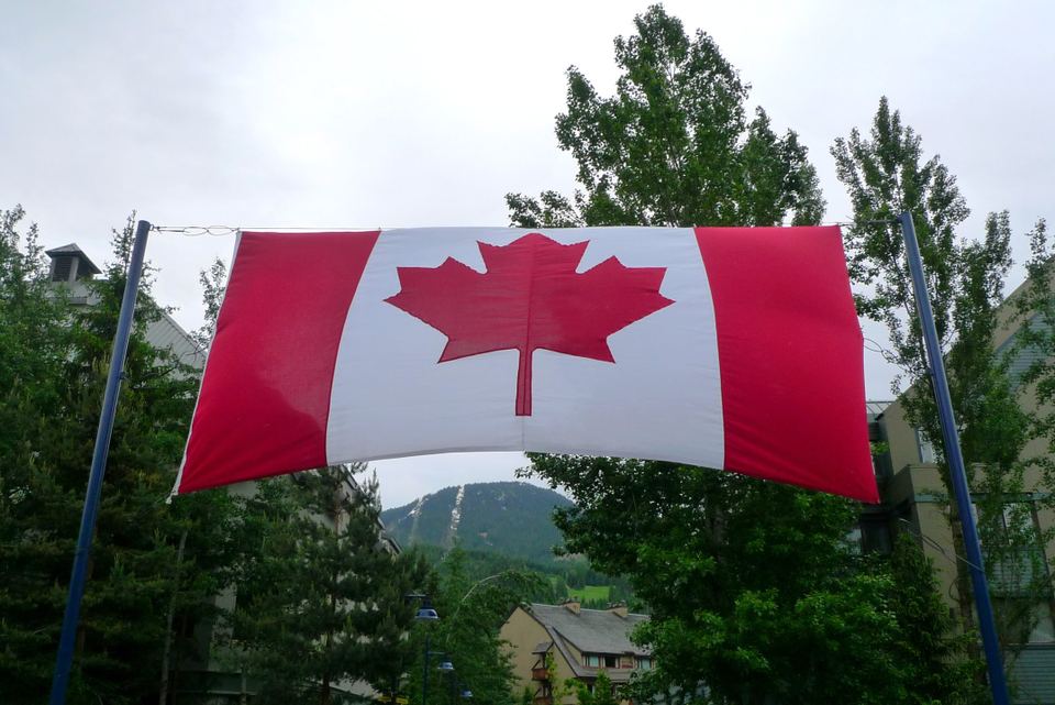 A large Canadian flag suspended in the air over green trees and alpine mountains.