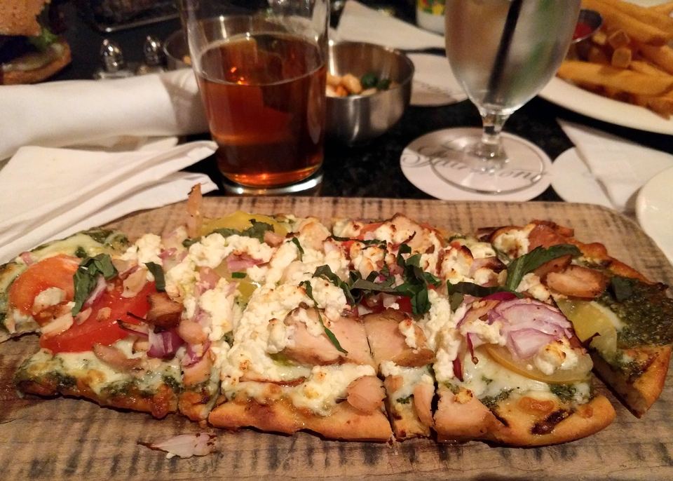 Flatbread topped with goat cheese and sliced tomatoes on a wooden board surrounded by drinks.