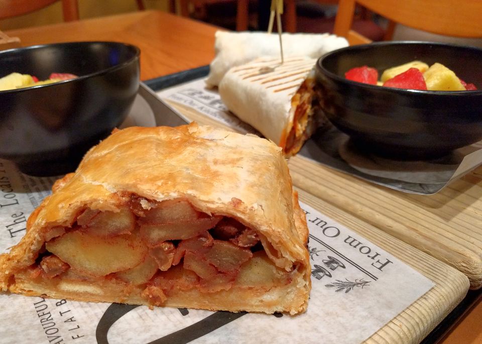 A slice of apple strudel pastry served on paper next to bowls of cut fruit.