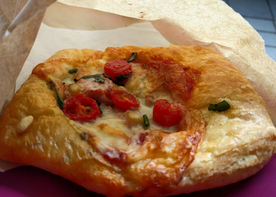 A close-up of a savory baked pastry topped with tomatoes and melted cheese inside a paper bag