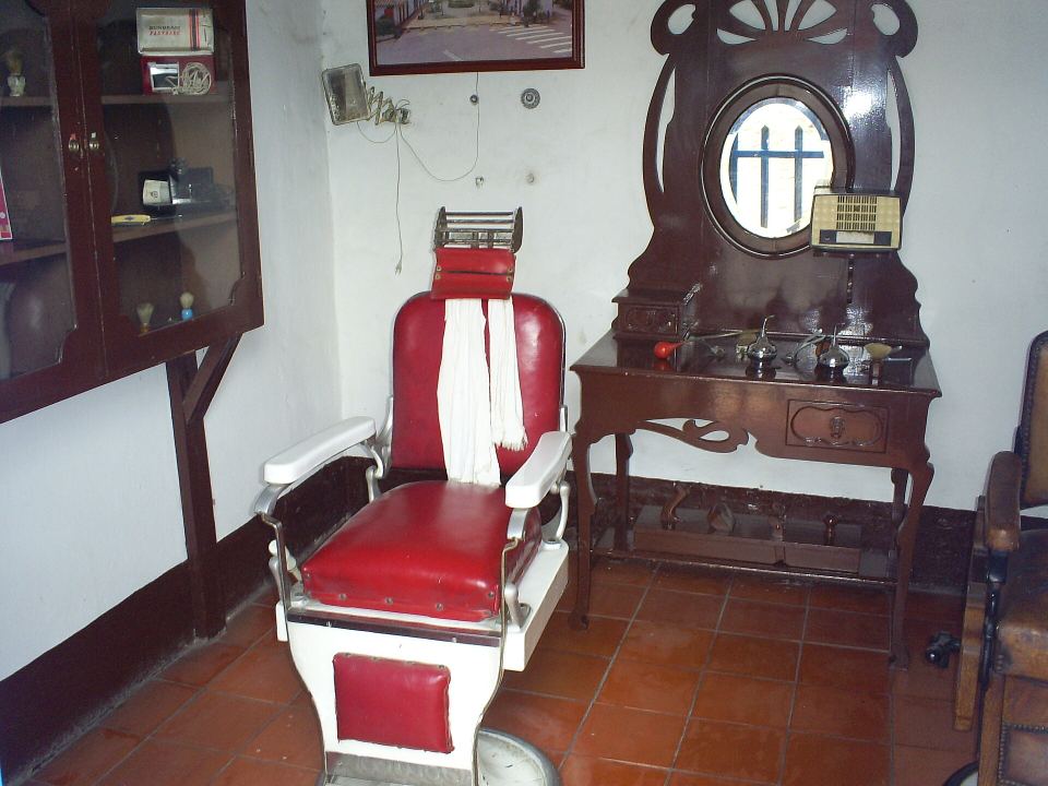 A vintage red-and-white barber chair beside a wooden vanity table and mirror in a white room.