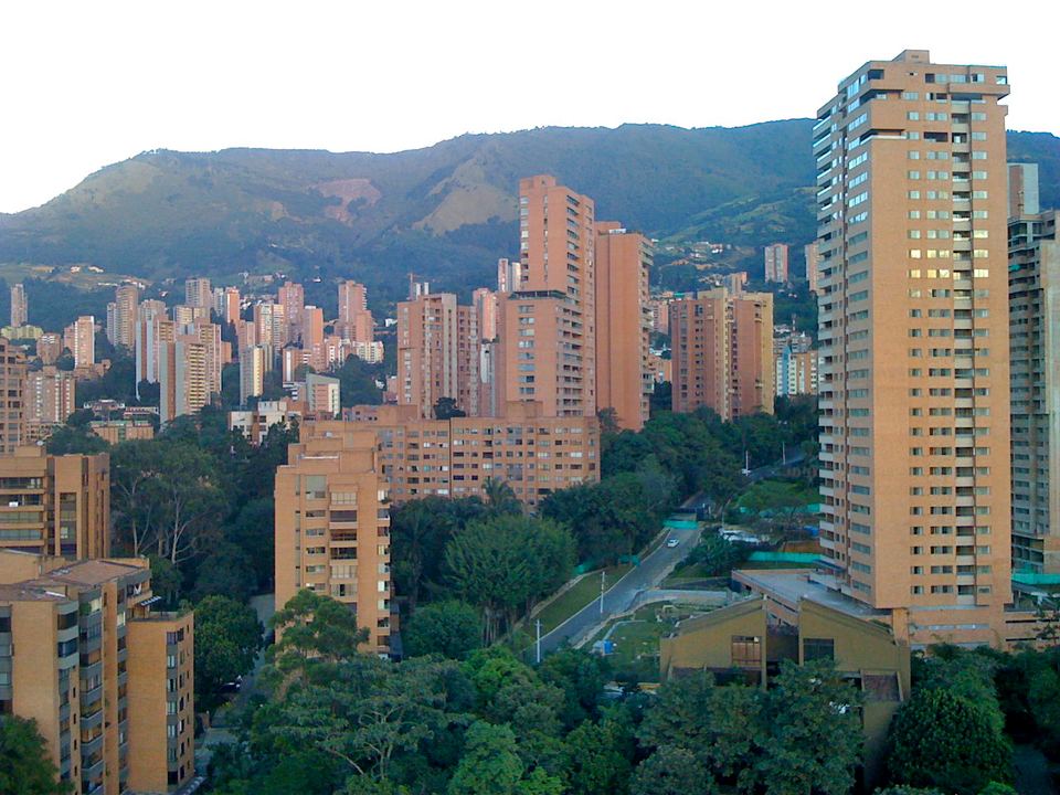 Tall brick residential towers surrounded by green trees in front of a blue mountain backdrop.