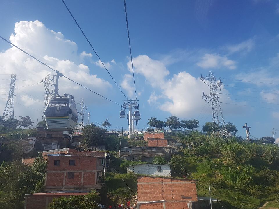 A white cable car hovering over brick houses on a green hillside against a blue sky with clouds.