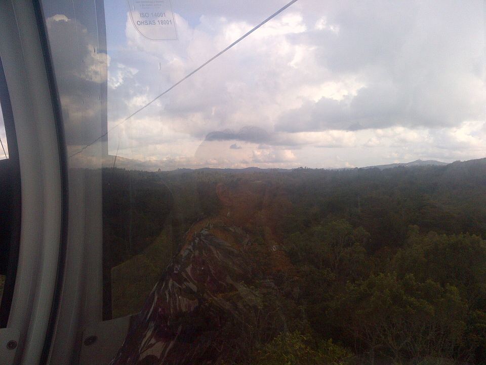 View through a cable car window looking down over a vast green forest canopy under cloudy skies.