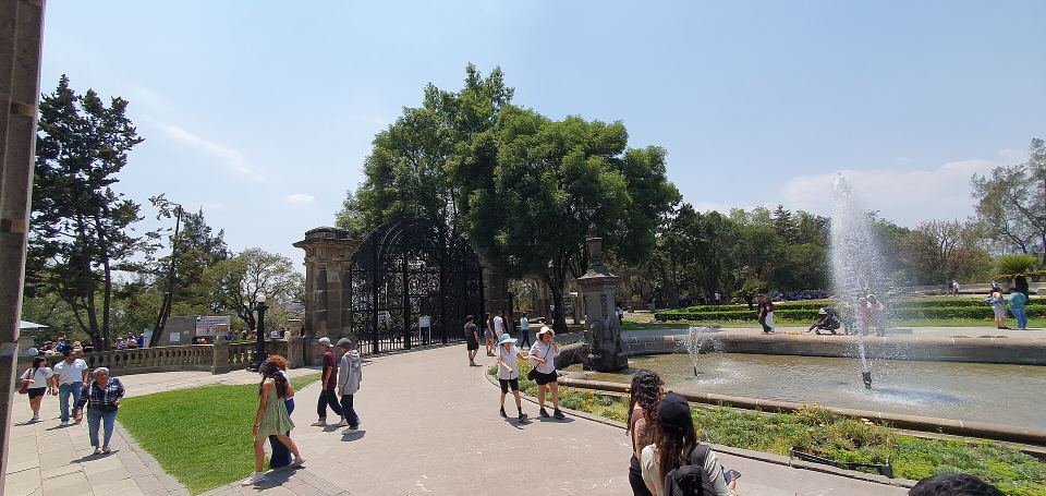 Pedestrians walking along a paved path past an ornate iron gate toward a fountain.