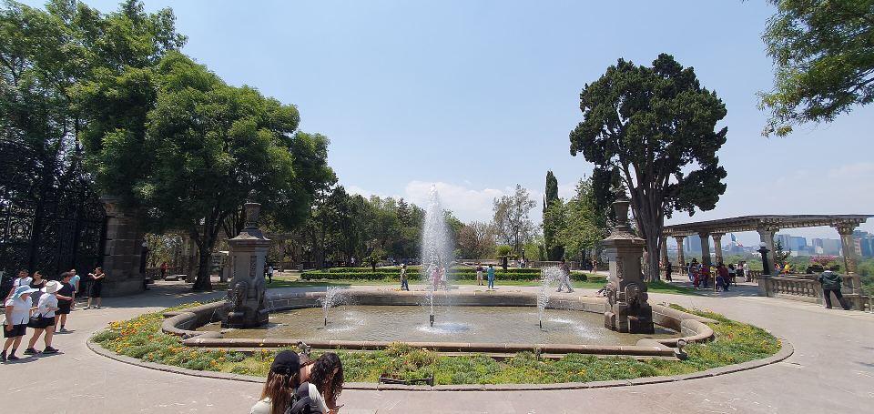 Wide view of a circular stone fountain spraying water in a leafy public park.