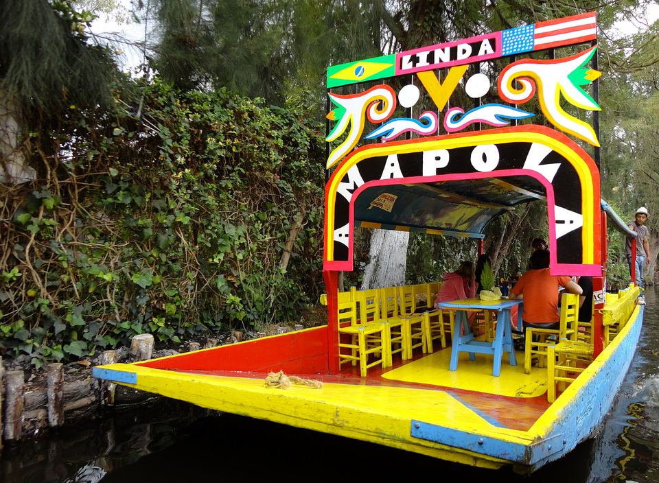 A bright yellow trajinera boat with passengers floating down a tree-lined canal.