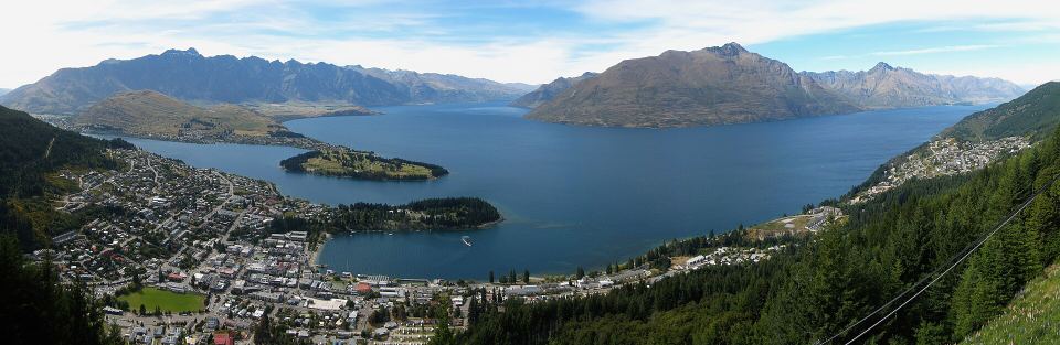 Panoramic aerial view of a lakeside town tucked between deep blue water and steep mountain ranges.