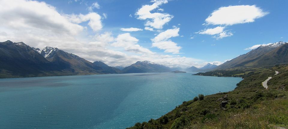 Winding road alongside a vast bright blue lake surrounded by snow-dusted mountains under a cloudy sky.