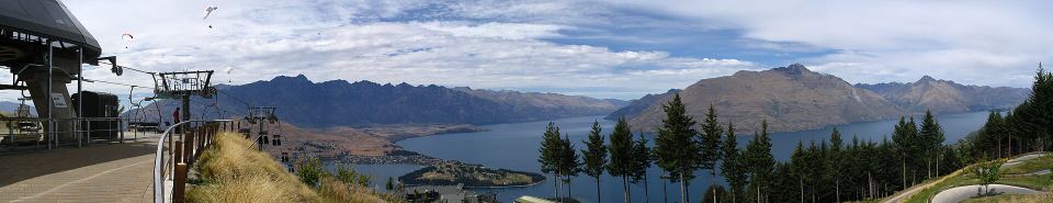 High mountain lookout showing a luge track, chairlift terminal, and paragliders floating over a lake.