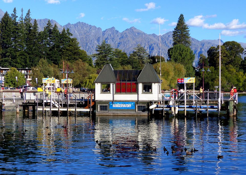 Small wooden observatory building on a lake pier with mountains in the background and ducks swimming.