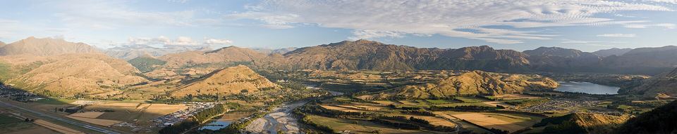 Panoramic high-angle view over golden farmland, a winding braided river, and mountain ridges.