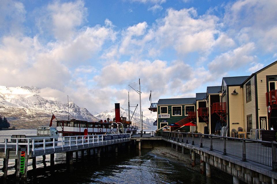 Historic steamship docked at a wooden pier alongside waterfront buildings and snow-capped mountains.