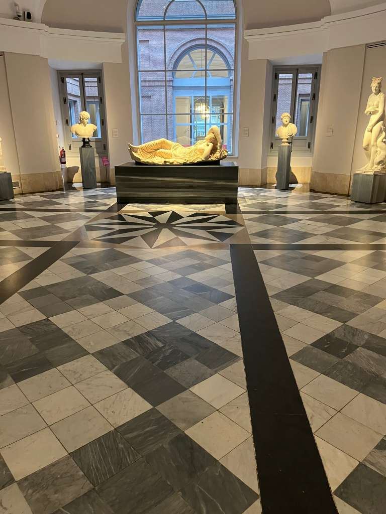 A marble reclining sculpture centered on a black and white checkered floor in a high-ceilinged museum.