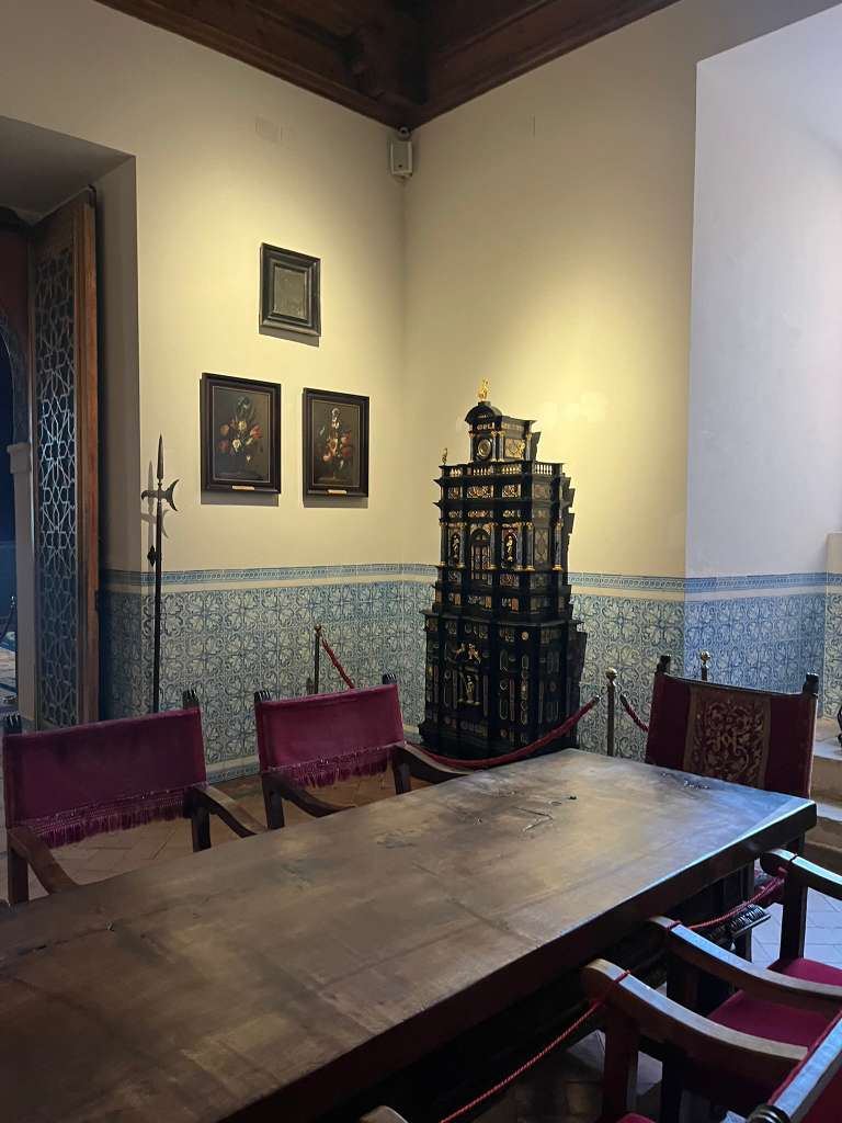 A long wooden table and red velvet chairs in a historic room with blue ceramic tile wall accents.