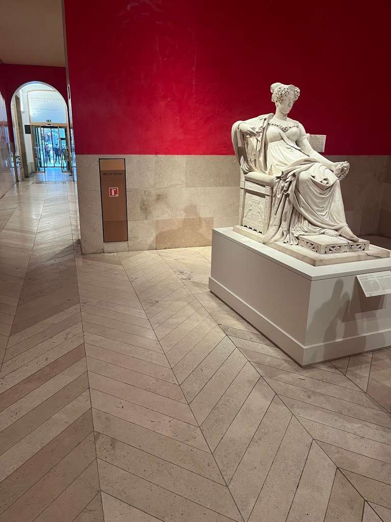 Marble sculpture of a seated draped woman displayed against a bright red gallery wall.