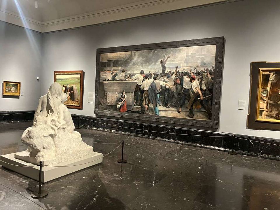 Large canvas painting of factory workers hung in a grey museum gallery beside sculptures.