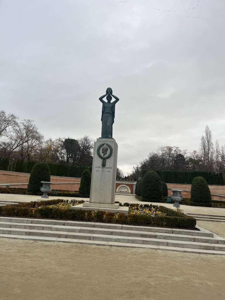 A bronze statue of a figure holding a mask atop a tall stone monument in a park garden.