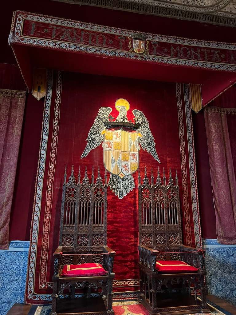 Two carved wooden throne chairs set against a red velvet wall displaying a royal emblem.