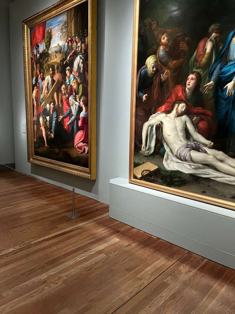 Large Renaissance paintings of religious scenes displayed on grey gallery walls above wood flooring.
