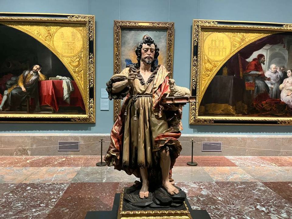 A carved wooden statue standing in an art museum gallery between large gold-framed paintings.