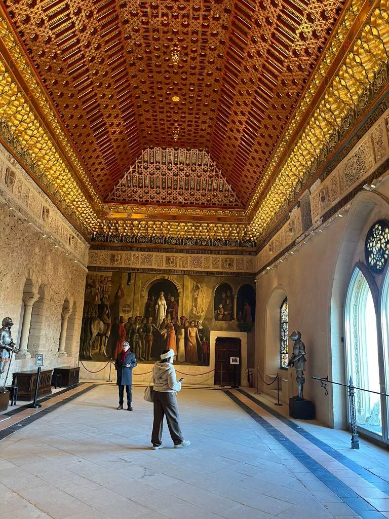 Visitors inside a castle hall with suits of armor and an ornate golden ceiling.