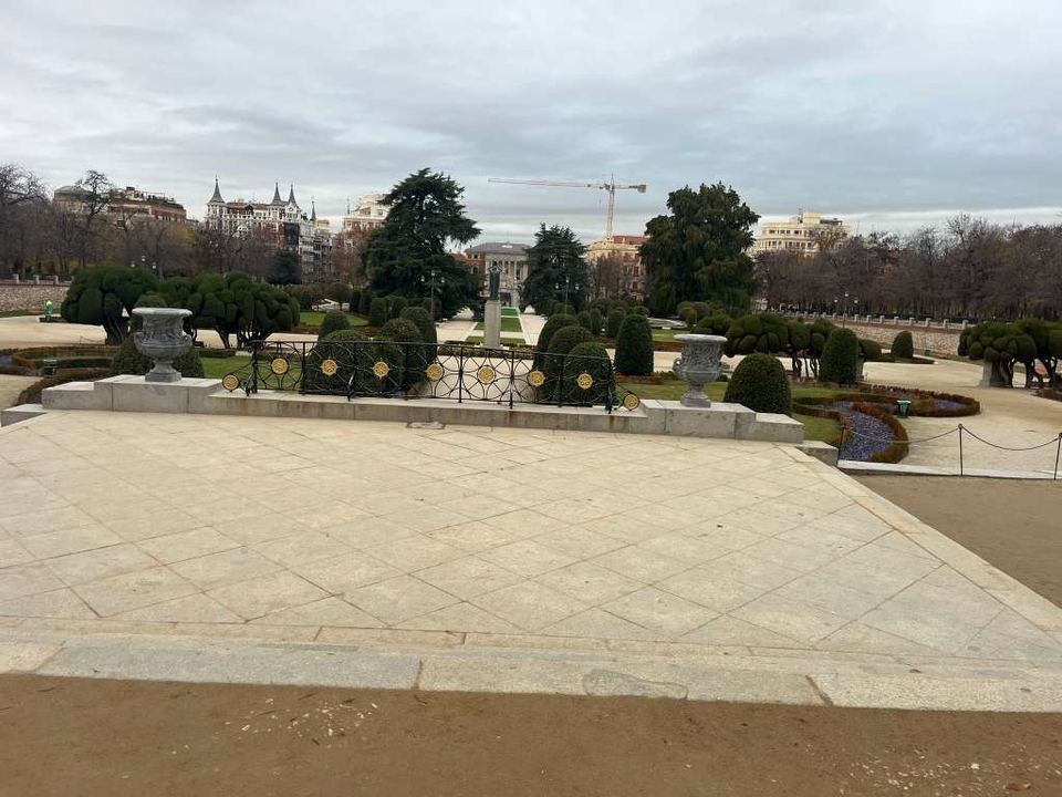 Manicured park garden with stone urns, topiary trees, and city buildings under an overcast sky.